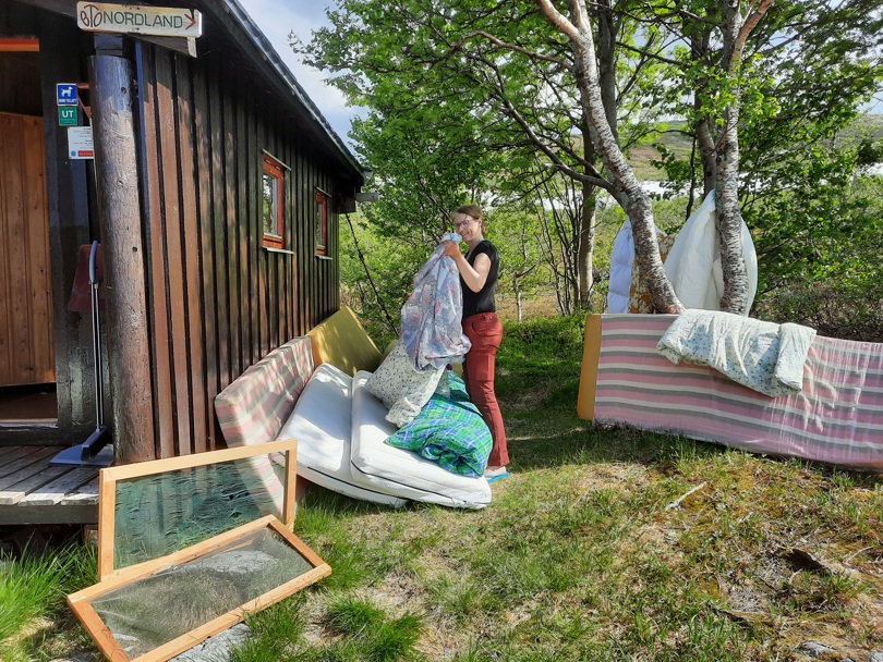 Ei dame står utenfor ei brunmalt hytte med sengetøy i hånda. Det ligger madrasser, myggnetting og mer sengetøy rundt omkring til lufting. Det er grønt og trær i bakgrunnen. Sol og pent vær.