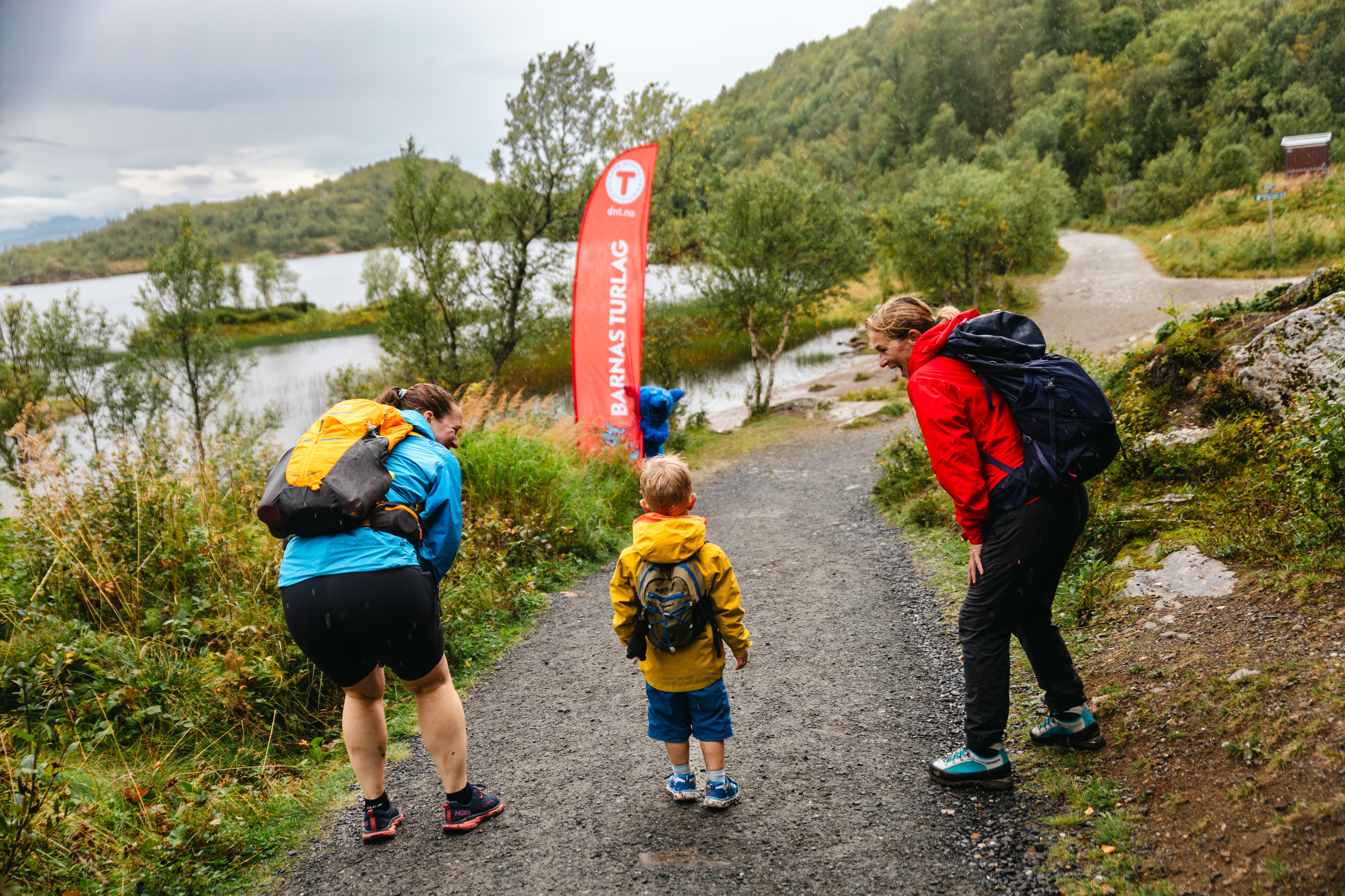 To damer og en liten gutt ser mot en blå maskot som gjemmer seg bak et strandflagg for Barnas Turlag