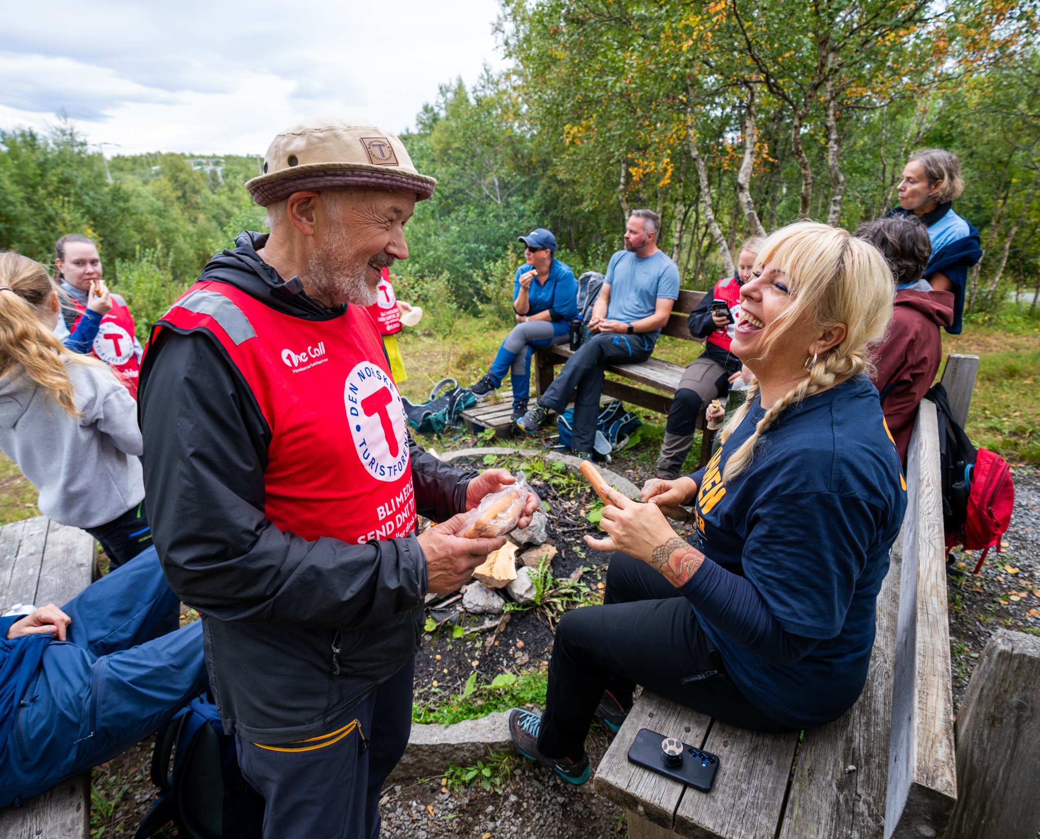 Mann med hatt og rød refleksvest serverer pølse til smilende dame med lyst hår