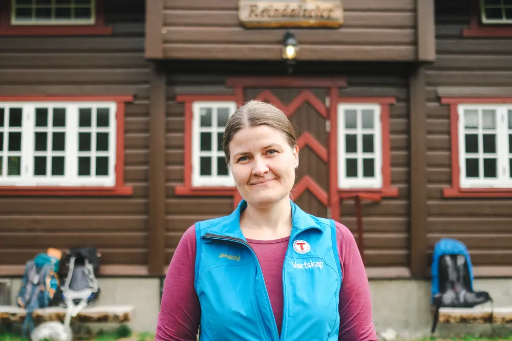 dame med blå vest med t på står på et hyttetun med et bredt smil om munnen