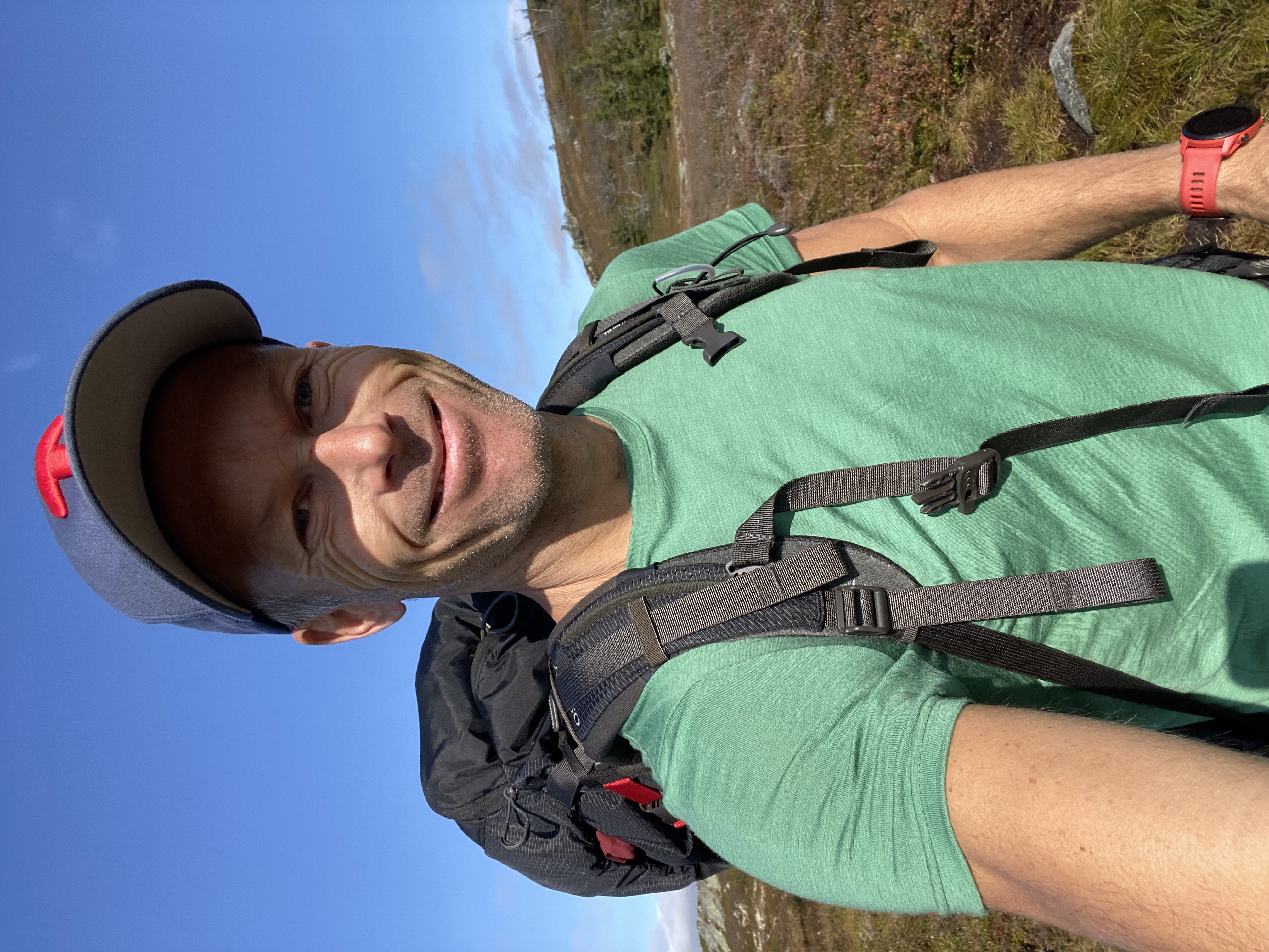 mann ikledd caps, grønn tskjorte og tursekk. smiler til kameraet og har et fjellandsskap bak seg