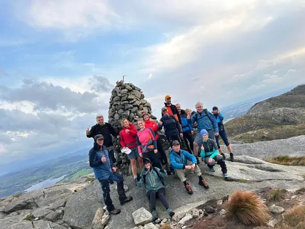 Gruppe med mennesker står foran varde på fjelltopp.