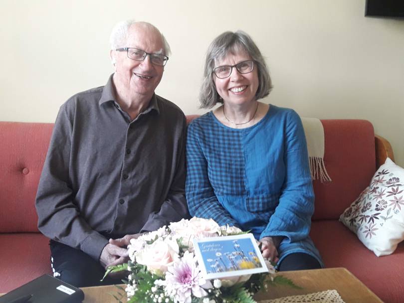 Eldre herre og litt yngre dame sitter i sofa. Fin blomsterbukett på bordet.