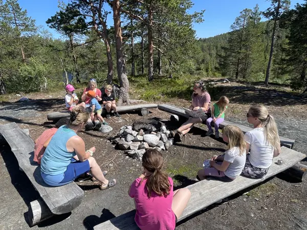 Voksne og barn sitter på benker rundt en bålplass