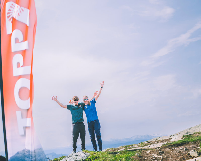 To personer står og veiver med armene på et fjell ved siden av et TOPP7-beachflagg.