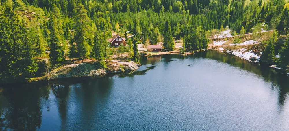 Dronebilde av vann og skog med hytte i bakgrunn