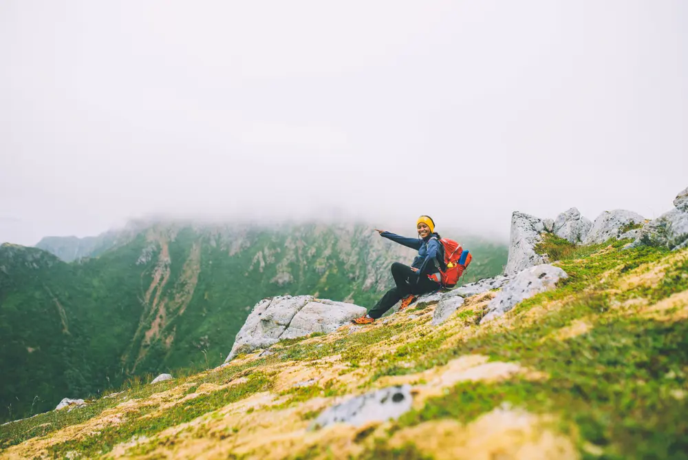 Kvinne sitter på en fjellside og peker, fjelltoppene rundt er dekt av tåke.