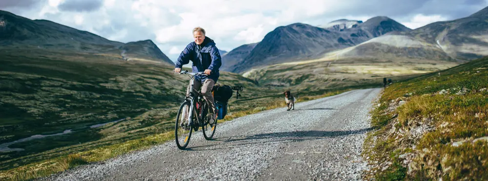 Syklist på grusvei med hund løpende bak i vakre fjellomgivelser. 
