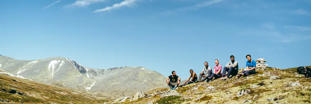 En gruppe mennesker sitter på en fjellrygg, men høye fjell i bakgrunnen.