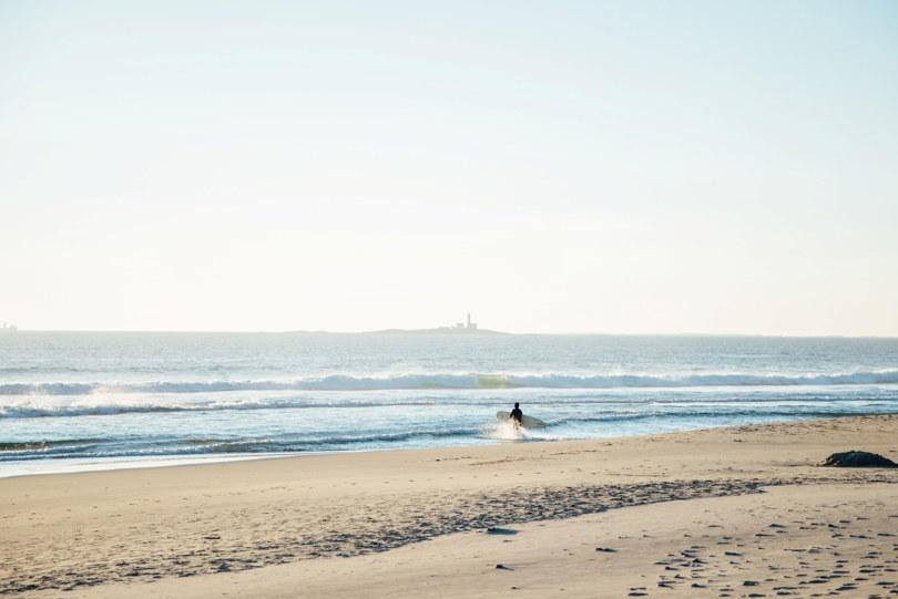 Ensom surfer på strand med høye bølger. Bak skimtes et fyr i horisonten.