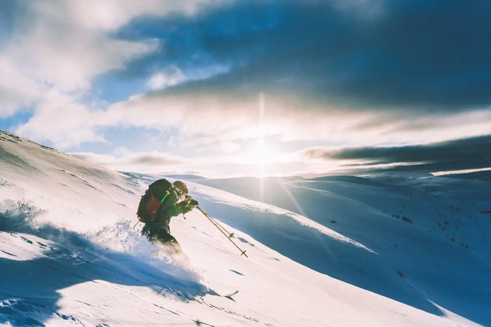 Person med vinterklær og tursekk som står på slalomski ned en snøkledd bakke, med solen som skinner gjennom skyene i bakgrunnen. 