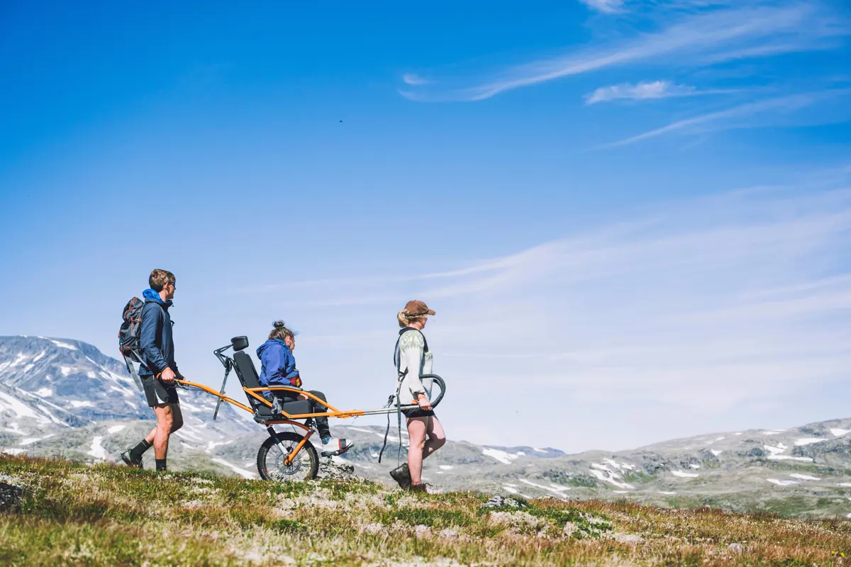 Tre personer ute på tur, på rekke og rad, sett fra siden idet de reiser fra venste mot høyre. Den midterste sitter i en rullestol. Noen fjell i det fjerne. Skyfri himmel. 