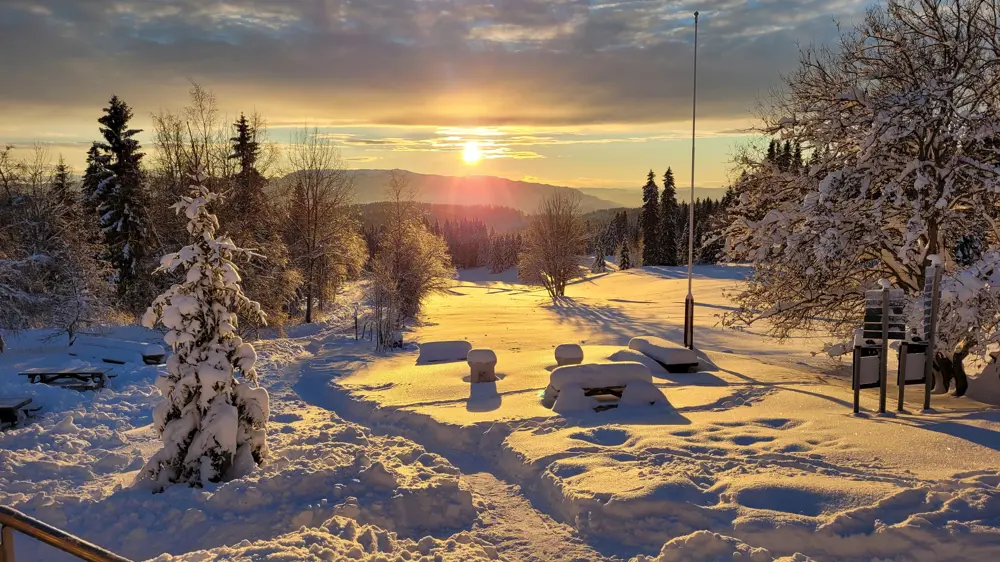 Landskap dekket med tykt lag nysnø, både benker og skilt nesten helt gjemt under det. Trær rundt den åpne flaten. En lav sol henger rett over horisonten. 