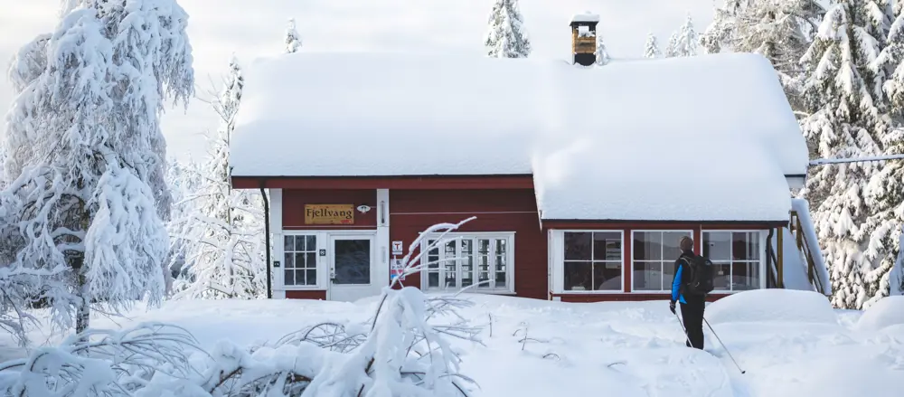 Mann foran en rødmalt hytte med mye snø på taket 