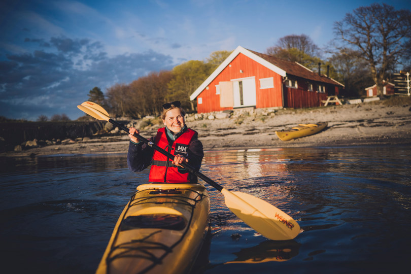 Dame i kajakk på fjorden. Rød hytte i bakgrunnen.