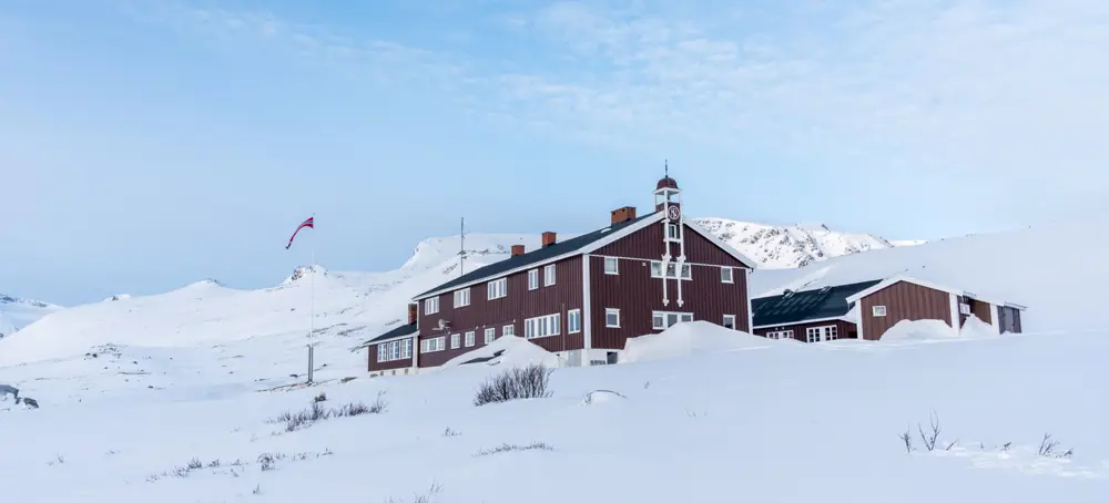 Rødbrune Glitterhiemhytta med klokkespir og flaggstang står i kontrast til den hvite fjellveggen. 