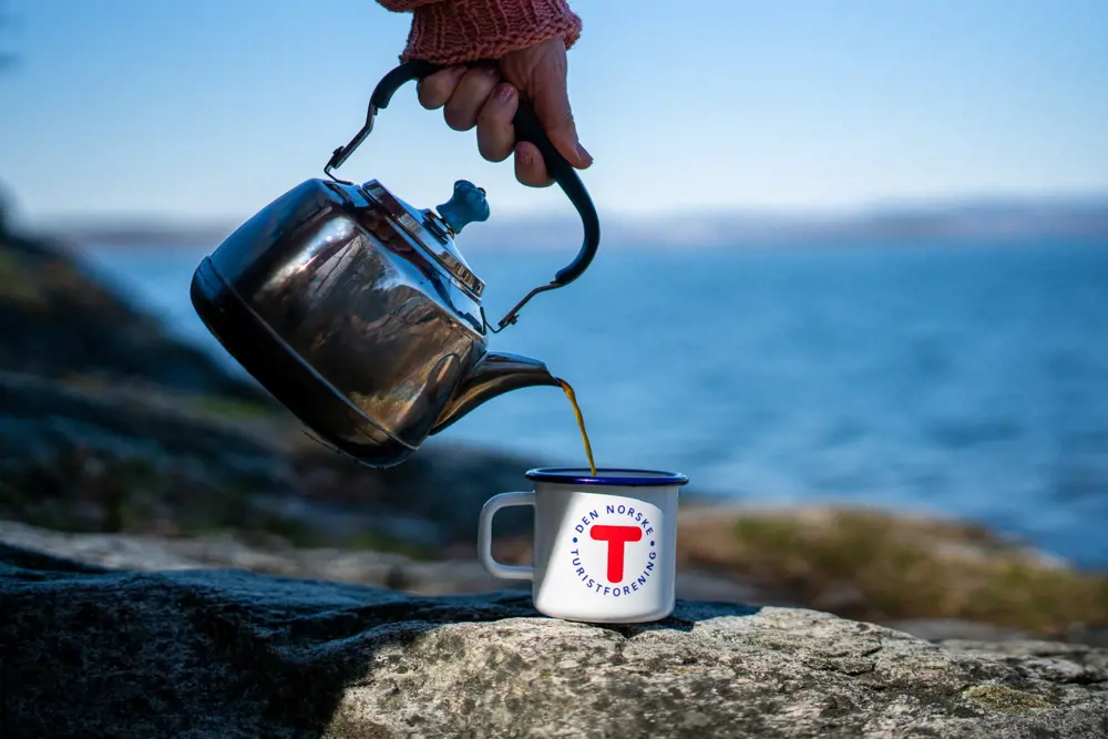 Noen heller fra en kaffekjele over i en hvit kopp med T-logoen på, med fjorden i bakgrunnen