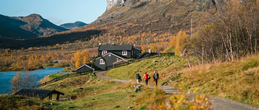 Tre turgåere, omgitt av spreke høstfarger, på grusveien inn mot porten foran den svarte hytta Gjendebu.