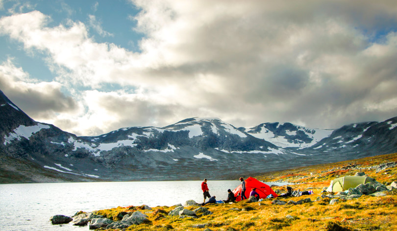 To telt ved et vann i mektig fjellområde. Noen mennesker rundt ved teltplassen.