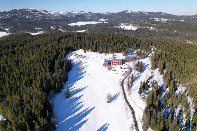 Dronebilde av en rød gård omgitt av granskog. Det er vinter og snø.