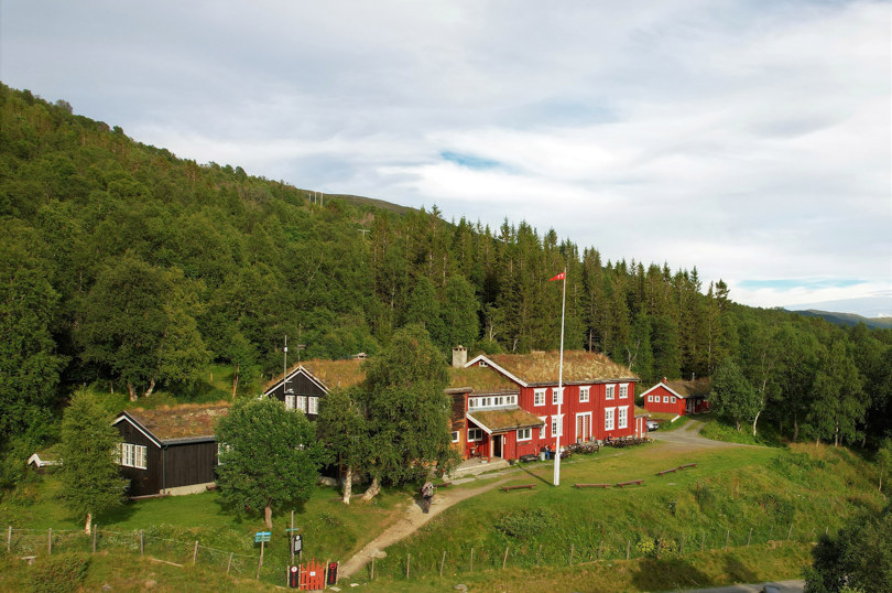 En hytte, rød på høyresiden, brun på venstre siden, med et par andre bygg rundt, med en skogdekket bakke bar, og en sti som leder fra frontdøren og mot kamera. 