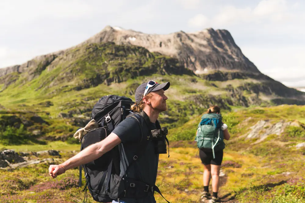 To personer ute på tur, begge med stor tursekk på ryggen. Stort fjell i mellomgrunnen