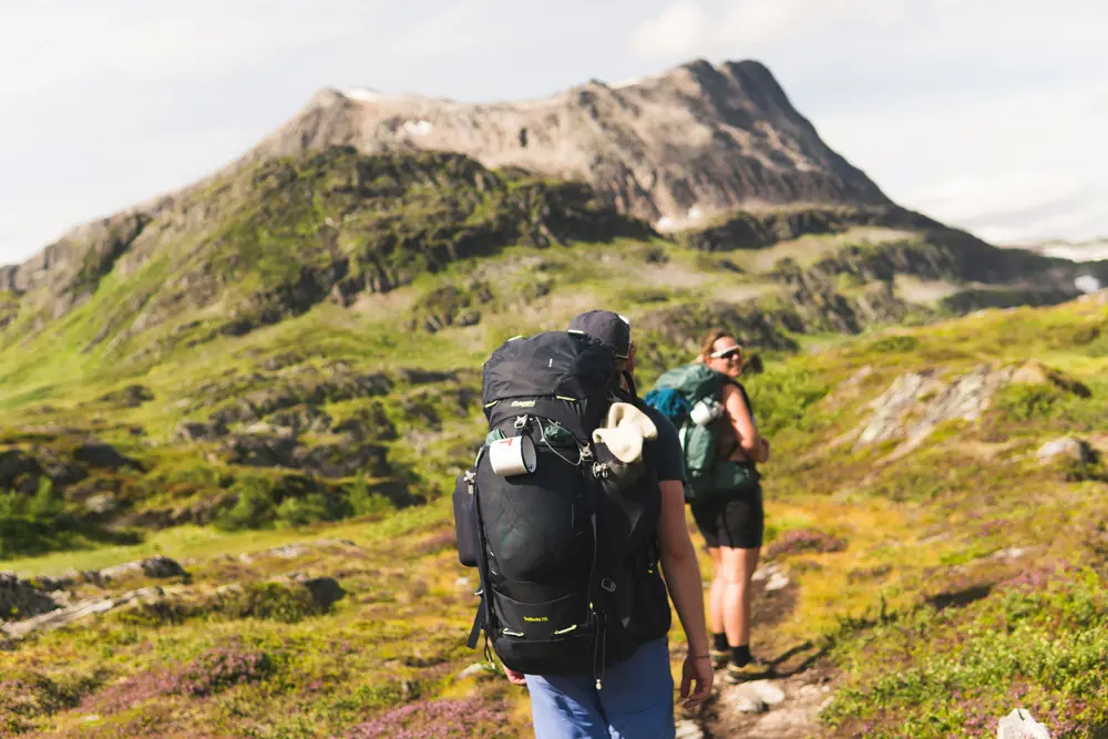 Person med stor tursekk sett bakfra, foran et villmarkslandskap med et fjell i bakgrunnen