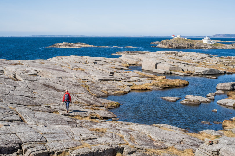 Kvinne i rød jakke vandrer på svaberg med havet og et fyr i bakgrunnen.