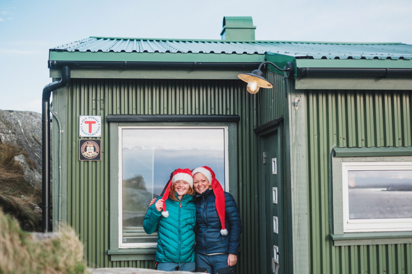 To damer med nisselue står foran inngangsdøra til ei grønn hytte med rød T på.