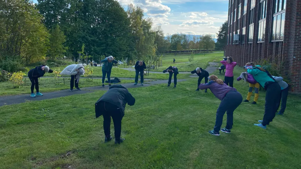 En gjeng med folk i treningstøy som står i en stor sirkel i en park, og bøyer seg og trener