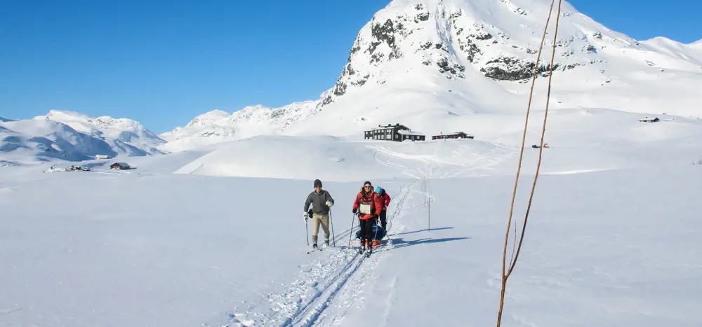 Tre personer som går på fjellski med en svart hytte i bakgrunnen.
