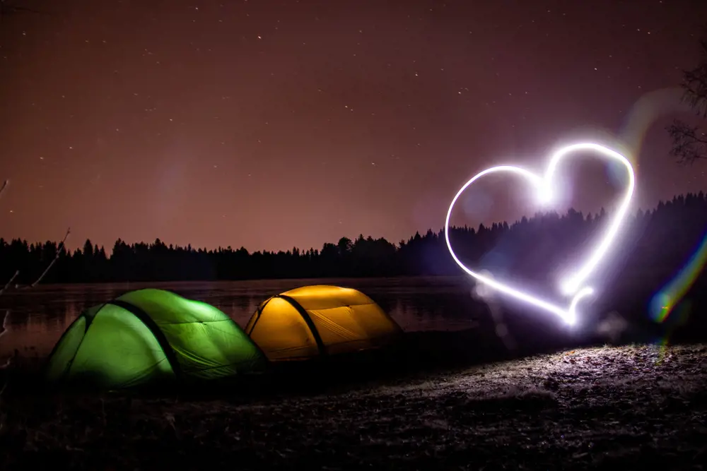 To telt lyser opp i forskjellige farger ved et tjern i skogen. Et stort hjerte laget av lys henger i lufta ved siden av teltene. Det er mørkt, og på himmelen ser man stjerner og lys etter et fly. 