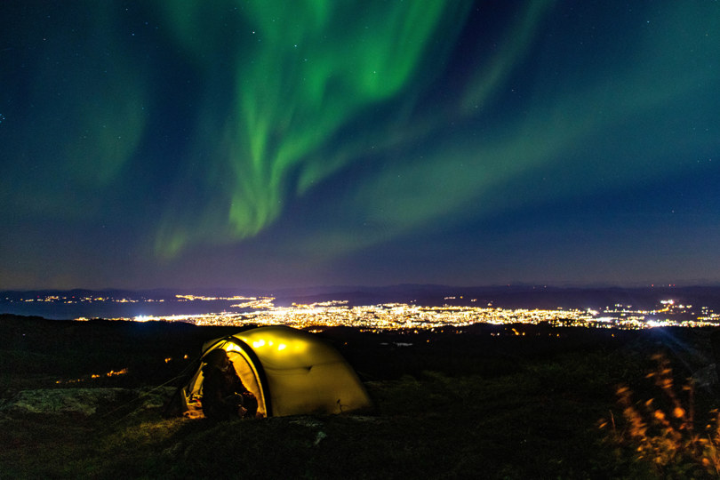 Bildet viser en mann ved et telt på toppen av et fjell, men utsikt over en stor by. Det er natt, og nordlys.