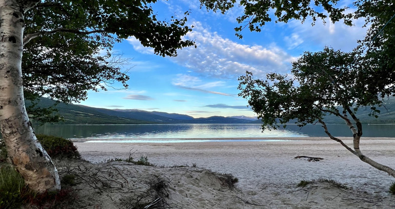 Bildet viser en stor sandstrand ved en innsjø omgitt av fjell.