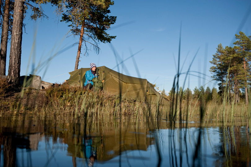 Et grønt telt i skogen ved et vann. En jente med blå jakke sitter i teltåpningen