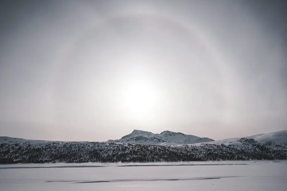  Bildet viser en stor åpen hvit himmel, med en blek sol i midten, omgitt av en ring-halo av lys, med et snødekket fjell rett under, som er del av en fjellkjede som strekker langs horisonten. Snødekket slette i forgrunnen.