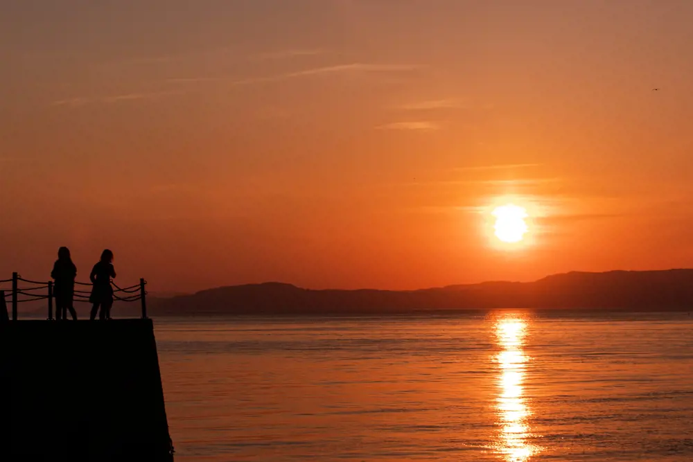 Bildet viser silhuetten av to figurer og kaia de står på i venstre bunn-hjørne, mens resten av bildet viser en solnedgang over fjorden, med landmassen på andre siden av fjorden synlig i det fjerne.