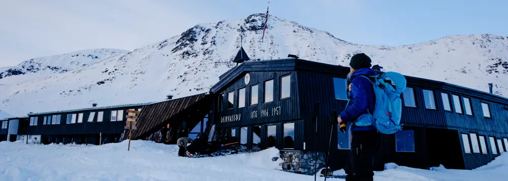 Svart hytte med stort T-merke og Leirvassbu markert på frontpartiet, en dame ser på hytta og fjellet i bakgrunnen. 