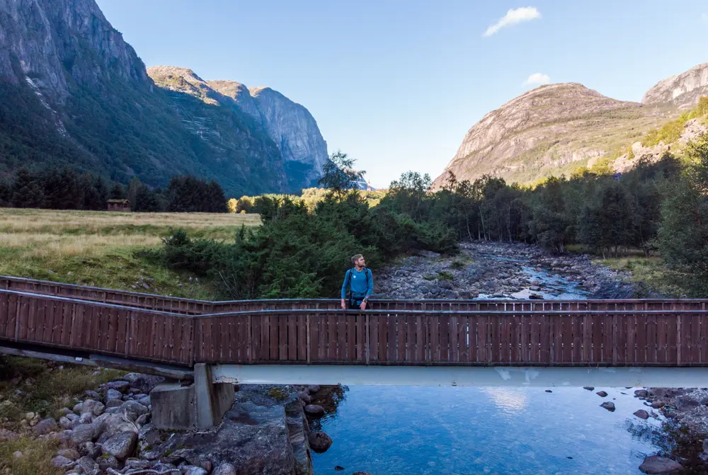 Bilde av en person som står på en bro som går over en elv. Morgenlys.