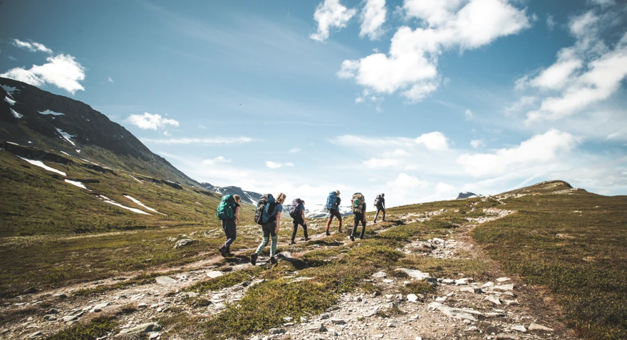 Bildet viser en gruppe mennesker på rekke og rad langs en sti i fjellet i fint sommervær.
