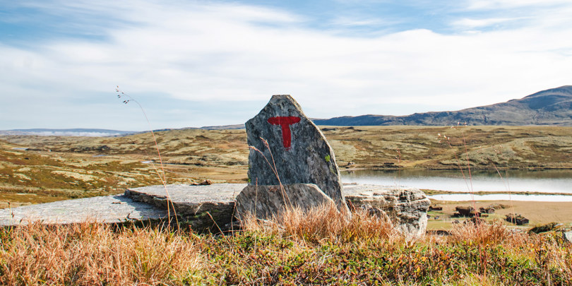 Bildet viser en varde med bokstaven T malt i rødt. Et høyfjellslandskap i tidlig høstfarger omkranser varden, og det er sollys og blå himmel.