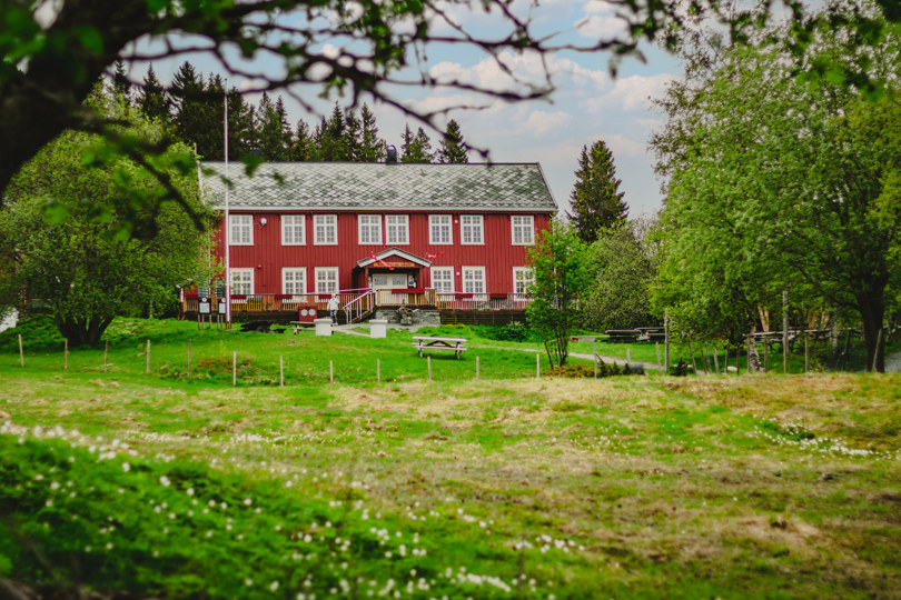 Et stort rødt gårdshus omgitt av skog, og en stor gresslette i forkant. Det er vår, og hvitveisen blomstrer.