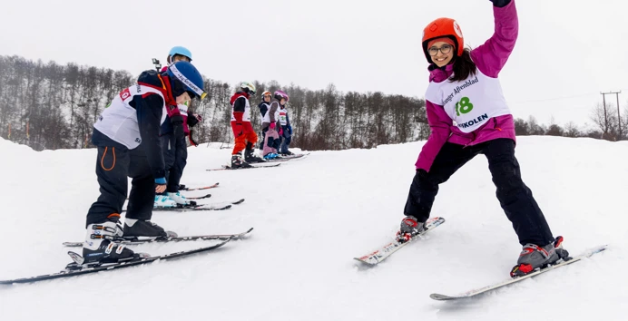 En jente i rosa jakke og skiskole-vest renner på slalom og løfter litt med den ene armen for å holde balansen.