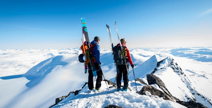 To personer som står på toppen av en fjelltopp med sekk og ski på ryggen