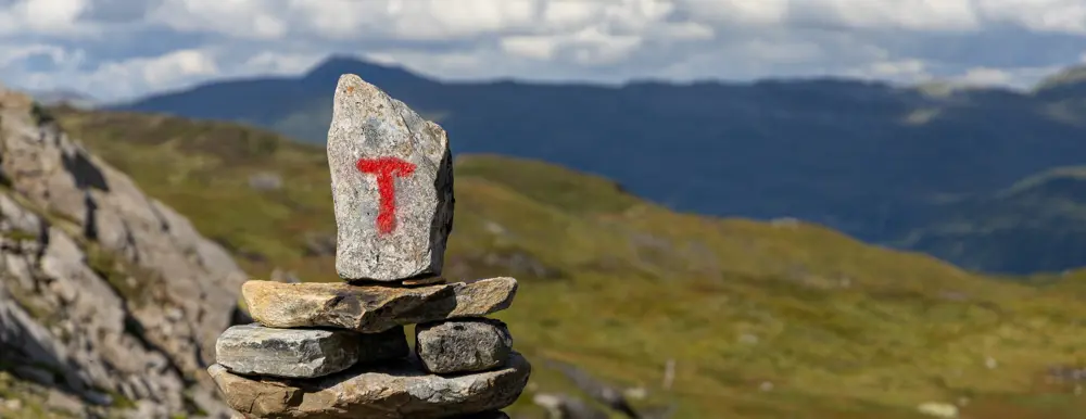 Rødmalt T på varde med fjell og lette skyer i bakgrunnen.