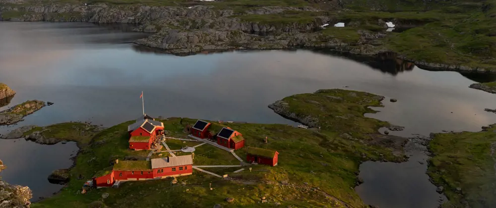 Dronebilde av hytta med oversikt over hytta, vannet og fjellene.