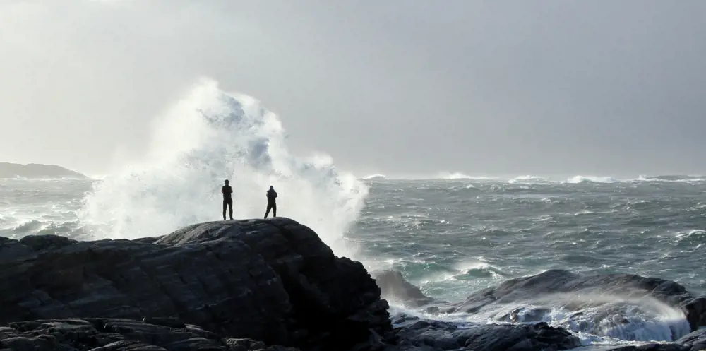 To mennesket står på svaberg og ser på store bølger ute i havgapet. Foto 