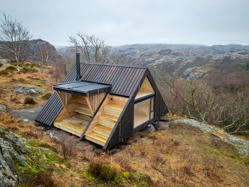 Oversiktsbilde av en brun trekantet hytte som ligger plassert i terrenget på en høyde.