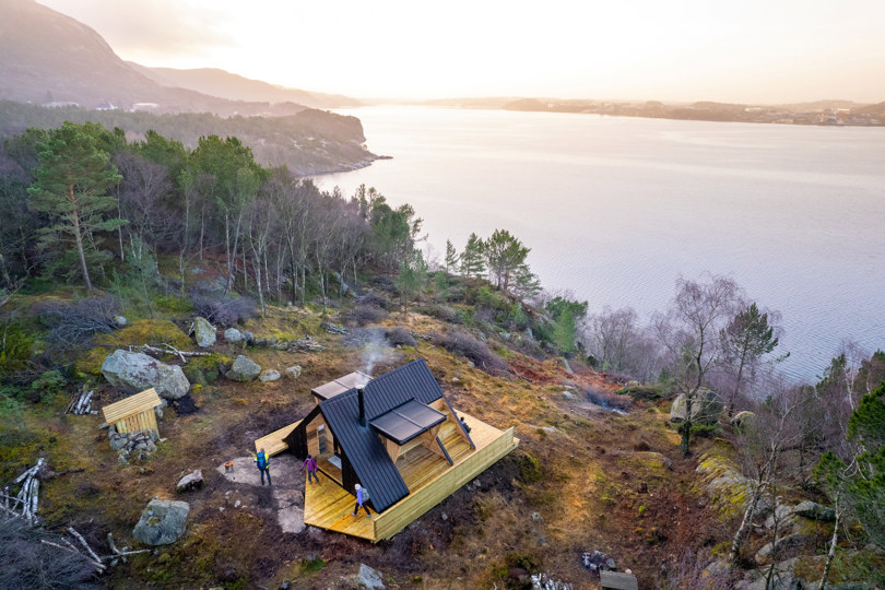 Dagsturhytte som ligger på toppen av et fjell med en lang fjord i bakgrunnen. Røyk fra pipa på hytta. Oversiktsbilde som er tatt med drone.