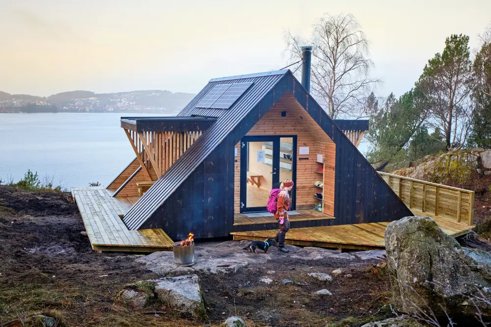 En liten hytte som ligger på toppen av et fjell med utsikt ut over fjorden. En kvinne med oransje genser og rosa ryggsekk går forbi inngangen med en hund i bånd.